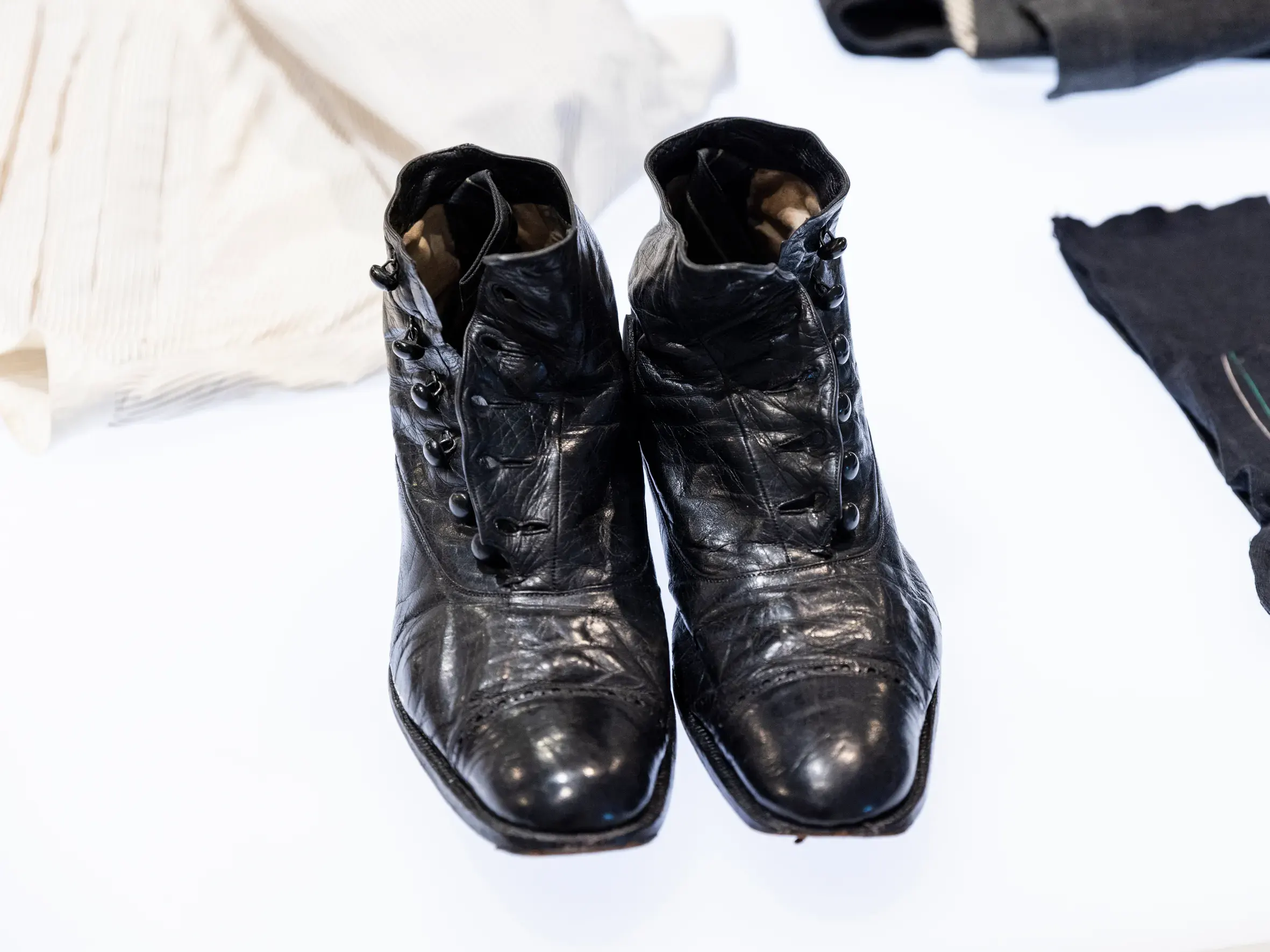 In der Mitte des Bildes steht ein Paar schwarz-glänzende Schuhe. Es handelt sich um eine Art Stiefeletten, die mit sechs Knöpfen zugemacht werden. Rechts davon liegen zwei dunkle Kleidungsstücke, von denen nur ein Teil zu sehen ist und auch das nur verschwommen. Links hinter den Schuhen liegt ein weisses Hemd mit schwarzen Streifen.